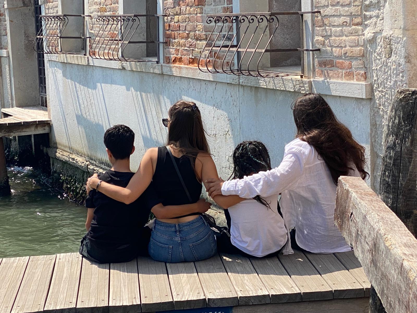Four children sitting arm in arm on a dock, backs turned, watching the water